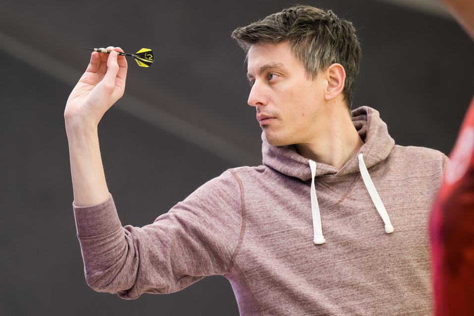 Ein Mann in einem grauen Hoodie zielt mit einem Dart auf eine Dartscheibe in einer Turnierumgebung.