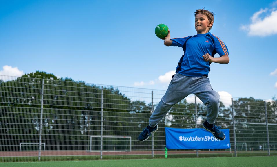 Junge springt auf einem Sportplatz und wirft einen grünen Handball, umgeben von einem Zaun und Bäumen im Hintergrund.